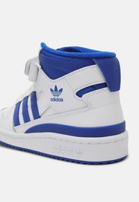 Valkoiset ja siniset korkeareunaiset Adidas-tennarit, joissa on kolme sinistä raitaa, Velcro-hihna, teksturoituja yksityiskohtia ja pehmustettu nilkkareuna.