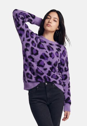 Frau trägt einen flauschigen violetten Pullover mit Leopardenmuster und schwarzen Flecken sowie schwarze Hose, posiert mit einem Arm hinter dem Kopf erhoben.