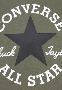 Olívaf green t-shirt nagy fekete csillag mintával, fehér szöveggel, amelyen a "Converse" és az "All Star" feliratok olvashatók vastag és kurzív betűtípussal.