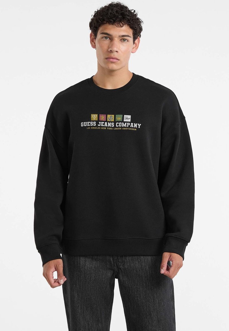 Młody mężczyzna z kręconymi włosami, ubrany w czarny sweatshirt marki Guess Jeans Company i ciemne dżinsy, stojący na jednolitym białym tle.