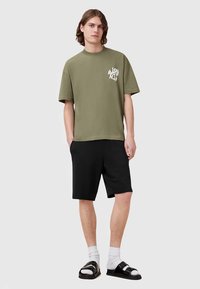 Olivově zelené tričko s krátkými rukávy a grafickým potiskem na hrudi, doplněné černými šortkami a černými sandály s bílými ponožkami.