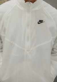 Vit Nike-jacka med hög krage, halvvindslås och nättextur. Har en liten svart logotyp på bröstet och sydda sömmar.