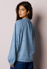 Mėlyna džinsinė marškinėliai su juodos širdies siuvinėjimu, sagomis užsegama apykakle, ilgomis rankovėmis su suspaustomis manžetėmis ir laisvo fasono.