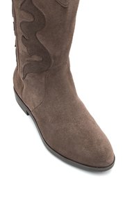 Bota de ante marrón hasta la rodilla con una textura suave, punta afilada y un patrón ondulado decorativo a lo largo del costado. Suela de goma plana en la parte inferior.