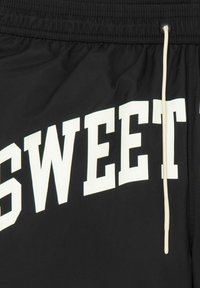 Un short noir en matériau léger présente un grand texte blanc "SWEET". Il est doté d'une taille élastique avec un cordon de serrage réglable.