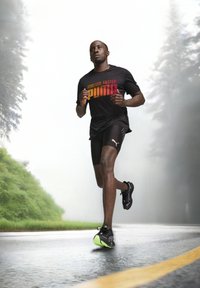 Crna sportska majica s natpisom "FOREVER. FASTER. PUMA" u gradijentnim bojama, uparen s crnim šortsevima i crnim tenisicama s neonsko zelenim detaljima.