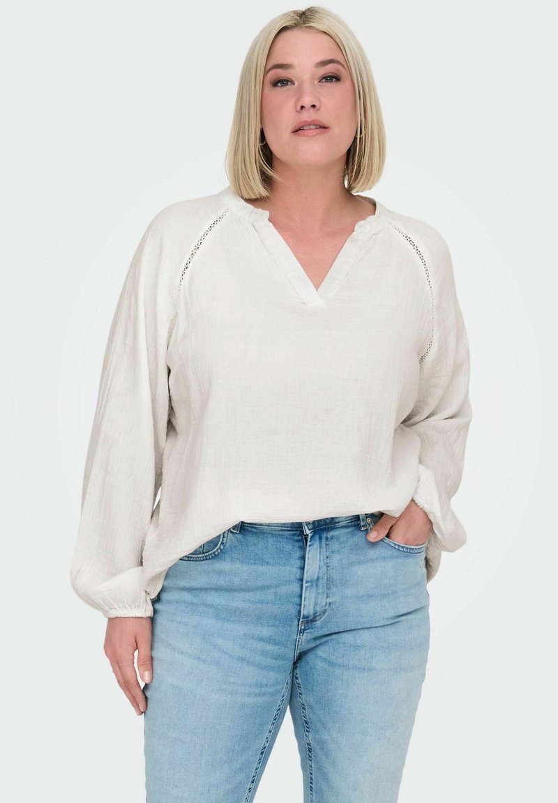 Blouse légère couleur beige clair avec un col en V, des manches longues et des détails en dentelle aux épaules, assortie à un jean en denim bleu clair.