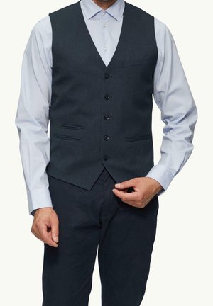 Mężczyzna ubrany w jasnoniebieską koszulę i ciemnoszary zapinany na guziki kamizelkę, poprawiający dół kamizelki, stojący na jednolitym tle.