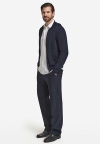 Marinblå huvtröja med en slät textur över en vit skjorta med knappar, kombinerad med mörka byxor och svarta loafers. Skräddarsydd, avslappnad passform.