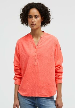 Woman with curly hair wearing a coral long-sleeve textured blouse and blue jeans, standing against a plain light background.