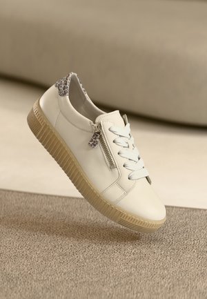 White leather sneaker with beige rubber sole, flat laces, side zipper, and leopard print detail on the heel. Textured fabric background.