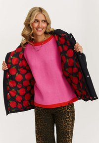 Žena v růžovém svetru a kalhotách s leopardím vzorem, která drží otevřenou černou prošívanou bundu s podšívkou v designu červených jablek.