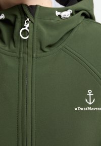 Veste softshell verte avec fermeture éclair à l'avant, capuche ajustable, et logo d'ancre blanc. Caractéristiques des coutures contrastées et une texture lisse.