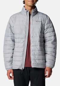 Helle graue isolierte Jacke mit einem gesteppte Design, hohem Kragen, durchgehendem Reißverschluss und Columbia-Logo auf der linken Brust, getragen über einem rötlichen Hemd.