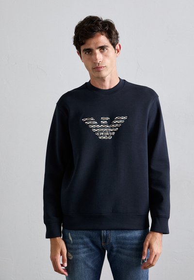 Mand bærer en sort sweatshirt med et mønstret ørn-logo og "GA" initialer, parret med blå slidte jeans, stående mod en ensfarvet væg.