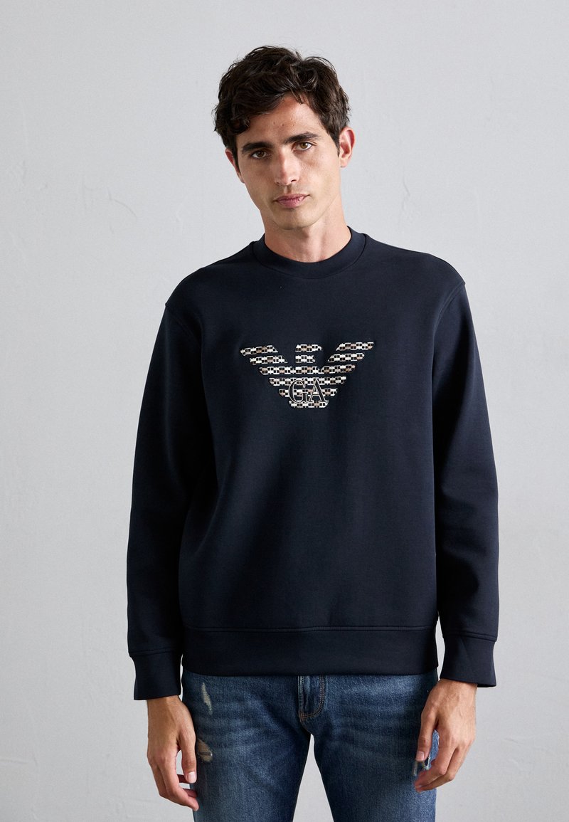 Man in een zwart sweatshirt met een patronen arend logo en de initialen "GA", gecombineerd met blauwe versleten jeans, staande tegen een effen muur.