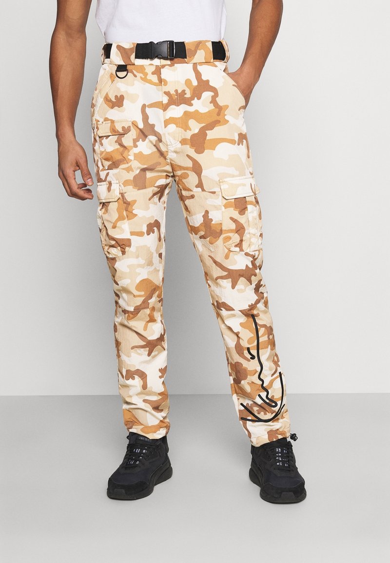 Homme portant un pantalon cargo camouflage beige et marron avec une ceinture noire et des baskets noires, debout devant un fond uni.