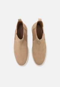 Botas de tobillo de ante beige con paneles laterales elásticos y tiradores, con una parte superior texturizada y una suela de goma blanca en contraste.