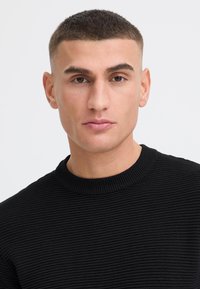 Jeune homme aux cheveux courts et foncés portant un pull noir côtelé, face à l'objectif avec une expression neutre sur un fond clair uni.