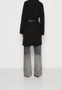 Svart kappa med bälte i midjan, tillsammans med vida grå jeans med en gråskala. Båda plaggen har en strukturerad design.