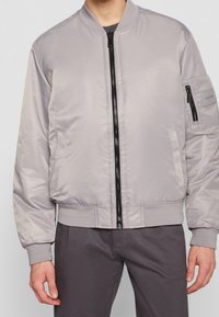 Giacca bomber grigia chiara realizzata in tessuto lucido, con zip frontale, colletto e polsini a costine, tasche laterali e una tasca utility sulla manica.