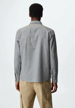 Mies, joka pukeutunut pitkähihaiseen sinivalkoiseen ruutupaitaan ja beigen sävyisiin työshortseihin, seisoo selin yksinkertaisen taustan edessä.