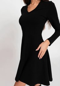 Robe noire à manches longues avec un col en V, un corsage ajusté et une jupe évasée. Confectionnée dans un tissu lisse avec une légère élasticité et sans motifs visibles.