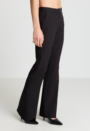 Schwarze, maßgeschneiderte Hose mit ausgestelltem Schnitt, ausgestattet mit einem flachen Bund, vorderen Falten und einer glatten Textur. Kombiniert mit schwarzen spitzen High Heels.