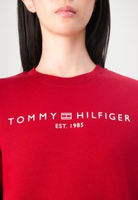 Červená mikina vyrobená z bavlny, s vroubkovaným výstřihem a bílým textovým logem "TOMMY HILFIGER EST. 1985" na hrudi.