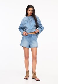 Jeanshuva med snörning och ficka, i kombination med ljusblå shorts med fransar. Tanfärgade, slappa sandaler avslutar outfiten.