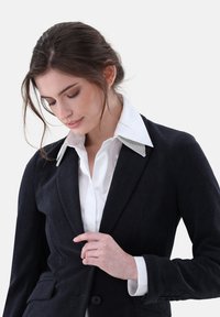 Marineblaue Cordjacke mit einem glatten weißen Hemd. Die Jacke zeichnet sich durch ein strukturiertes Design, eine taillierte Passform und einen mit Metallakzenten verzierten Kragen aus.