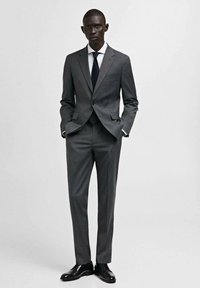 Completo grigio scuro composto da un blazer aderente e pantaloni su misura. L'outfit include una camicia bianca e una cravatta nera, completato da scarpe nere.