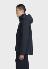 Námořnická modrá zateplená bunda s kapucí, s logem na rukávu, zipovým zapínáním a drukovými detaily. Texturovaná tkanina.