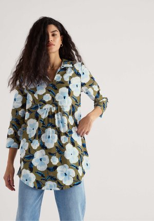 Žena s dlhými kučeravými vlasmi, oblečená v zelenej tunike s veľkými bielymi a modrými kvetmi a svetlomodrých džínsoch, stojaca pred neutrálnym pozadím.