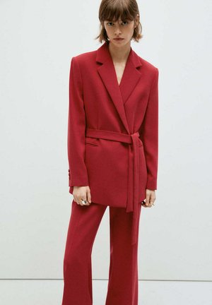 Young woman with short brown hair wearing a red belted blazer and matching wide-leg trousers, standing against a plain light background.