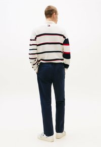 Pasiasty sweter w kolorach kremowym, granatowym i czerwonym, z długimi rękawami, połączony z granatowymi spodniami i białymi trampkami. Widoczny detal z logo na kołnierzyku.