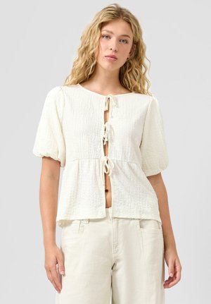 Młoda kobieta z długimi, falującymi blond włosami, ubrana w białą bluzkę o fakturze z trzema wiązaniami z przodu oraz jasne spodnie, na jednolitym tle.