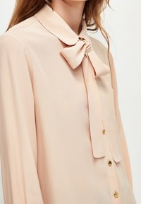 Blusa color pesca realizzata in tessuto leggero, con colletto rotondo, chiusura con fiocco e bottoni dorati lungo la parte anteriore.