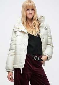 Dunjakke i kremfarge med høy krage, trykknapper og teksturert stoff. Brukt over en svart strikket topp og burgunderbukser.