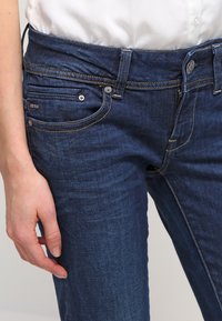 Dark blue denim jeans feature front pockets, subtle stitching details, and a mid-rise waistband secured with a metal button and zipper.