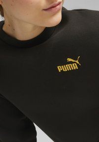 Mustmustrekiga musta värvi, ümarate kaelustega, millel on rindkeres silmapaistev kollane borduuritud Puma logo. Sile kangatekstuur.