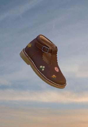 Chaussure enfant marron avec broderie florale et bride à boucle, sur un fond de ciel bleu doux.