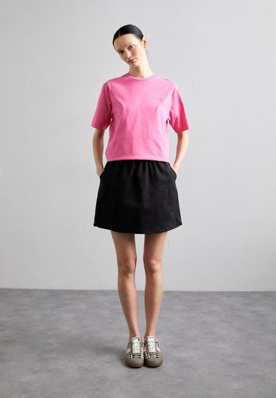 Rosa bomull t-skjorte med korte ermer, kombinert med et svart skjørt med elastisk midje. Modellen har på seg grå sneakers med mønstrede detaljer.