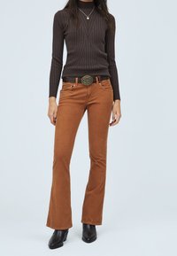 Braune Cord-Pumphosen mit glatter Textur, auf der Taille platziert, kombiniert mit einem dunkel gestreiften Langarmoberteil und schwarzen High-Heels-Stiefeln.