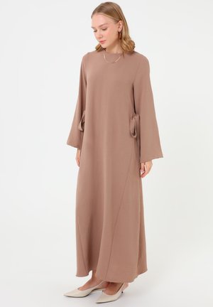Junge Frau trägt ein langes, lockeres braunes Kleid mit weiten Ärmeln und Taillenbändern, kombiniert mit beigen spitzen Ballerinas.
