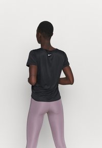 Person står med ryggen mot kameran, iklädd en svart Nike-t-shirt och mauve högmidjade leggings, mot en enkel vit bakgrund.