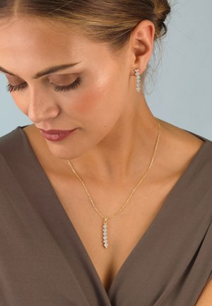 Goldene Halskette mit einem vertikalen Anhänger, der ein lineares Design aus kleinen, klaren Edelsteinen zeigt. Ergänzt durch passende goldene Ohrhänger.