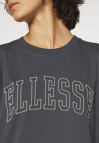 Camiseta de algodón gris oscuro con cuello redondo que presenta un gran logo en crema contrastante "ELLESSE" en el pecho.