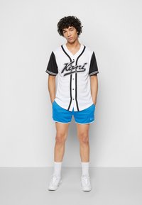 Hvit baseballtrøye med svarte ermer, svarte knapper og logo. Kombinert med blå shorts og hvite sneakers. Jevn stoffstruktur.