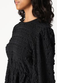 Vestido negro texturizado con un patrón de cuadrícula, mangas abullonadas y cuello redondeado. El material tiene un efecto tridimensional.